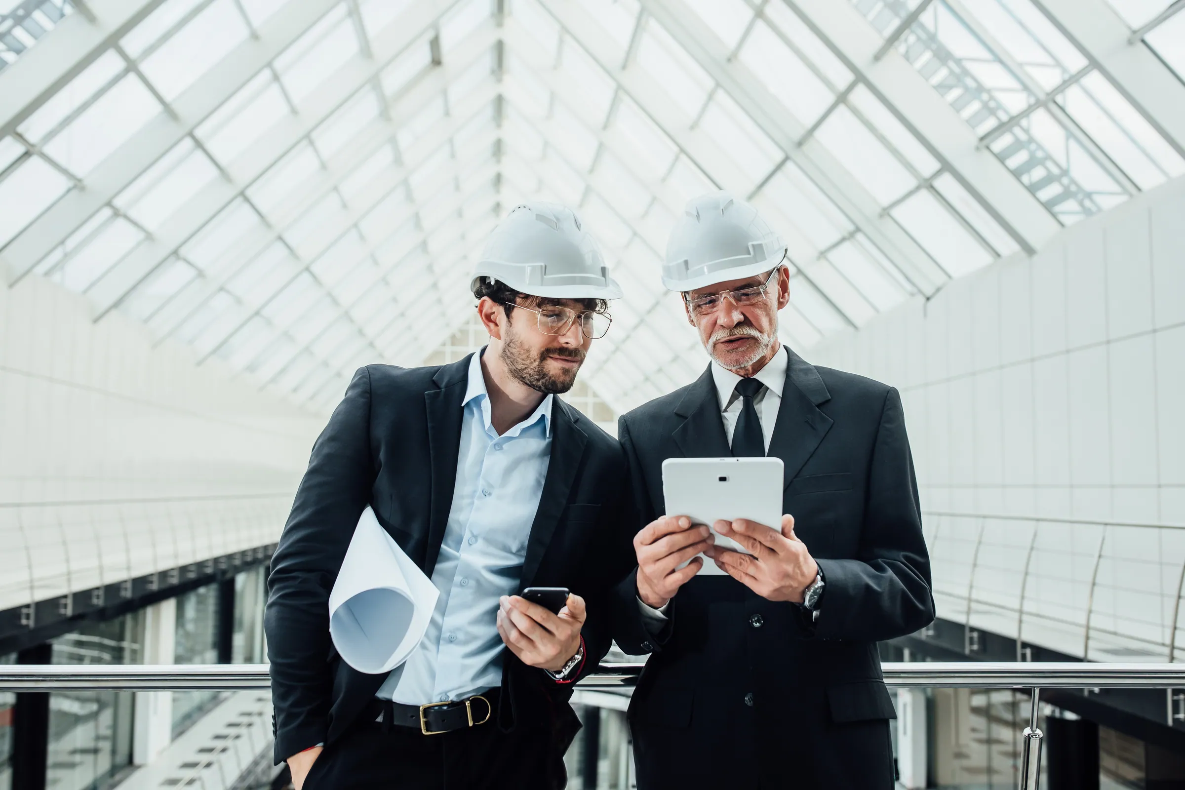Two managers in suits and hard hats with a tablet under a glass roof