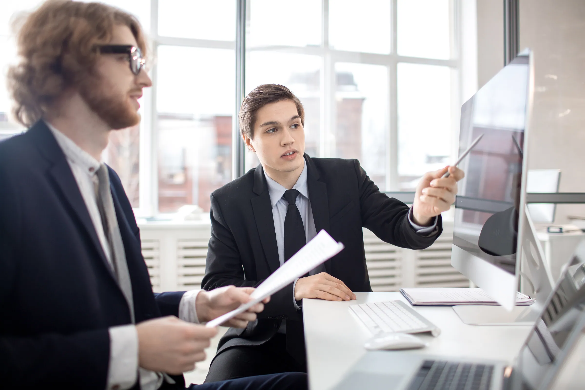 Consultants in suits reviewing work at a shared workstation, representing specialist talent backed by a practice
